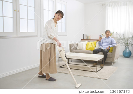 一位微笑的女士和一位六十多歲的男士一邊用吸塵器打掃一邊交談。 136567373