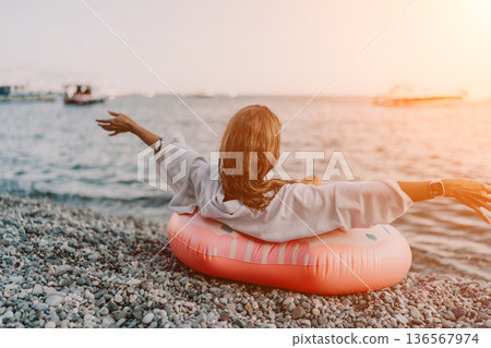 Woman, beach, ocean. Relaxing woman on pebble beach with inflatable ring, gazing at the beautiful sunset sea, copy space. 136567974