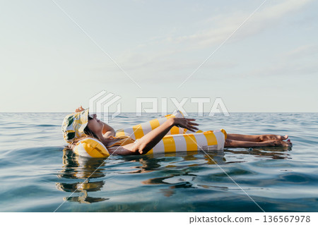 Woman Beach Float Relaxation: A woman wearing a straw hat floats on a yellow and white striped inflatable raft in the ocean. 136567978