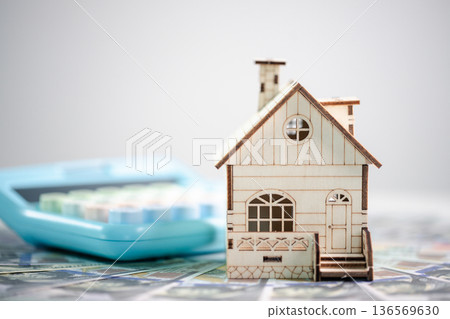 Miniature wooden house on scattered banknotes with blue calculator in the background on white, illustrating home price calculation, mortgage planning, long-term financial security. mortgage planning 136569630