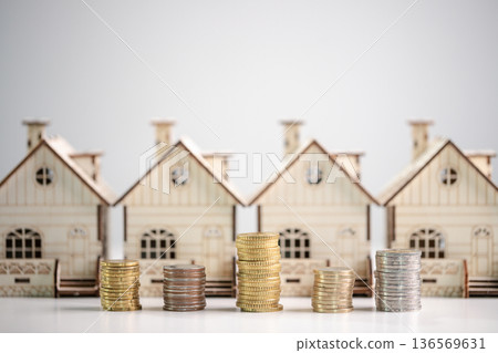 Wooden model houses behind stacks of coins on white surface, copy space, illustrating property value, rental income, mortgage rates, investment portfolio and long-term financial security. investment Wooden model houses behind stacks of coins on white surface, copy space, illustrating property value, rental income, mortgage rates, investment portfolio and long-term financial security. investment 136569631