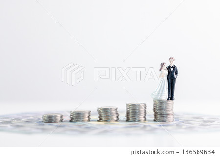 Bride and groom figurines standing on rising stacks of coins over banknotes with wide white copy space, suggesting wedding savings, budgeting and smart money management for financial growth. savings 136569634