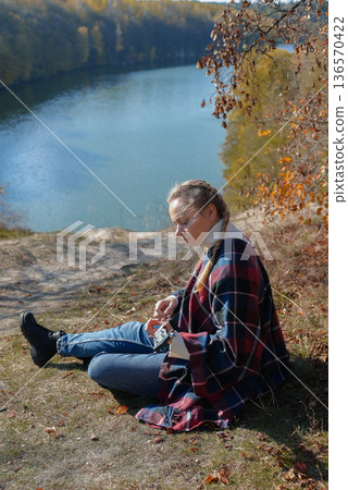Blonde woman playing guitar ukulele outdoors in autumn forest. Concept of sound therapy, mental health and wellness rituals. Calmness tranquility audio-sensory practices. Aura farming 136570422