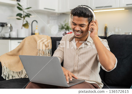 Indian man with wireless headphones edits short video on laptop works freelance smiles with approval 136572615