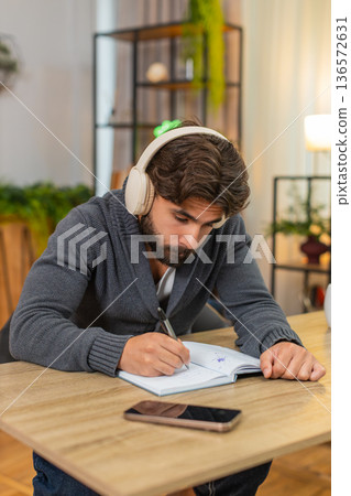 Indian man listens to audiobook on headphones takes notes in notebook focused intense study session 136572631