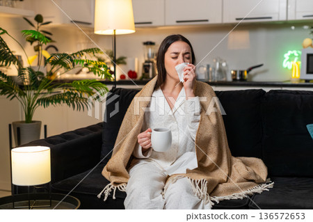 Young woman sick under blanket hot tea with meds sore throat tissues sneeze cough tired evening rest Young woman sick under blanket hot tea with meds sore throat tissues sneeze cough tired evening rest 136572653