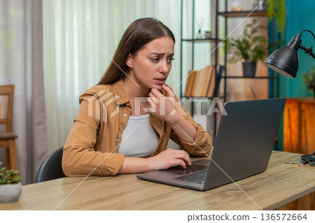 Young woman frowning at laptop reading worrying news showing stress and fatigue in late work session 136572664