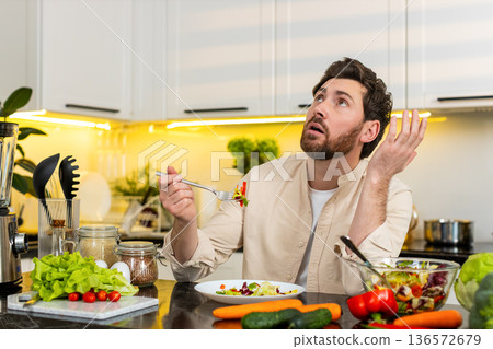 Middle-aged man unhappy eating fresh vegetable salad in home kitchen showing diet willpower struggle Middle-aged man unhappy eating fresh vegetable salad in home kitchen showing diet willpower struggle 136572679