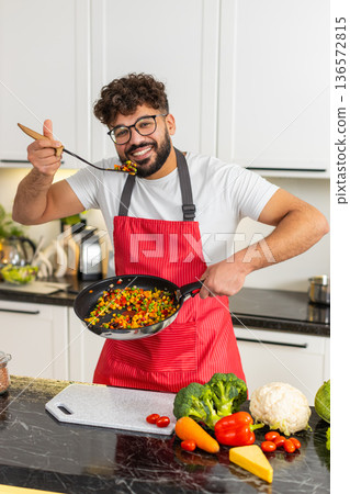 Arabian young adult man tasting vegetables from pan in kitchen cooking healthy dinner recipe at home Arabian young adult man tasting vegetables from pan in kitchen cooking healthy dinner recipe at home 136572815