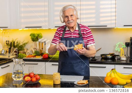Senior old man dancing with headphones while making healthy breakfast toast for diet in home kitchen 136572816