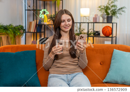 Young woman drinks water from glass plans tasks on smartphone focused calm and replies to messages Young woman drinks water from glass plans tasks on smartphone focused calm and replies to messages 136572909