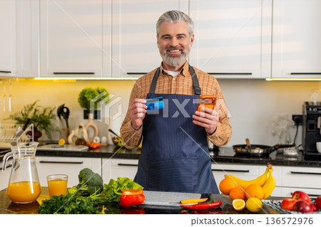 Middle-aged man uses smartphone with credit card in kitchen buying healthy groceries for diet plan 136572976
