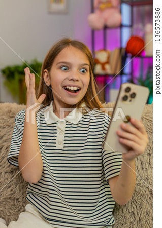 Child girl kid playing mobile game app on smartphone at home, smiling and celebrating happy victory Child girl kid playing mobile game app on smartphone at home, smiling and celebrating happy victory 136573886