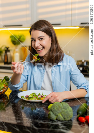 Young woman eating fresh salad in home kitchen enjoying healthy diet lunch for wellness routine joy Young woman eating fresh salad in home kitchen enjoying healthy diet lunch for wellness routine joy 136573893