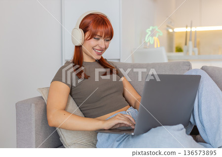 Young woman in headphones types email on laptop while music plays smiles and nods to rhythm focused 136573895