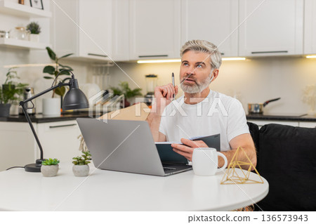 Middle-aged man studies online course with earphones watches lecture writes notes in notebook review Middle-aged man studies online course with earphones watches lecture writes notes in notebook review 136573943