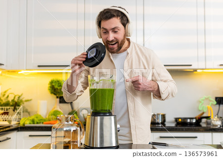 Middle-aged man excited wearing headphones showing green smoothie blender in home kitchen for diet Middle-aged man excited wearing headphones showing green smoothie blender in home kitchen for diet 136573961
