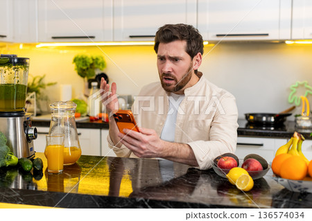 Middle-aged man shocked by smartphone alert in home kitchen reacting with surprise and worry moods 136574034