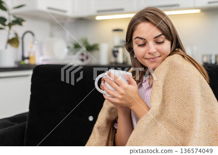 Young woman drinks herbal tea hugs warm mug snuggles in blanket smiles and enjoys peaceful cozy time Young woman drinks herbal tea hugs warm mug snuggles in blanket smiles and enjoys peaceful cozy time 136574049