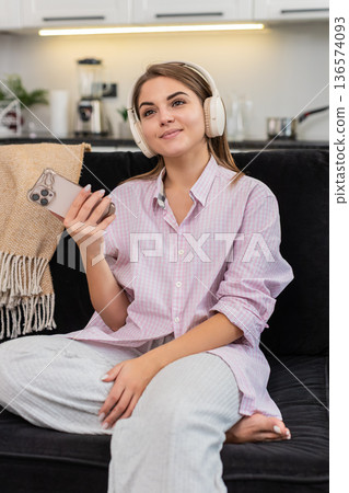 Young woman with wireless headphones nods to music writes idea in smartphone notes and chats online Young woman with wireless headphones nods to music writes idea in smartphone notes and chats online 136574093