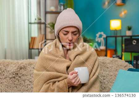 Young woman drinking hot tea in cozy blanket wearing hat feeling cold and sick resting on sofa today Young woman drinking hot tea in cozy blanket wearing hat feeling cold and sick resting on sofa today 136574130