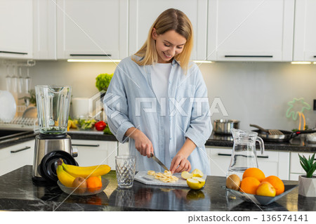 Young woman making smoothie adding banana apple spinach spirulina to blender detox in home kitchen 136574141