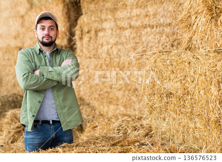 Male farmer posing against the background of hay bales in hayloft Male farmer posing against the background of hay bales in hayloft 136576523