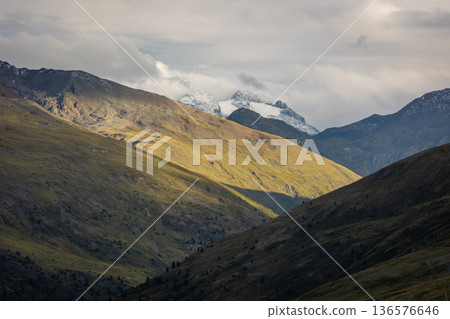 Mountain valley illuminated by soft sunlight with rolling alpine slopes and distant snow-covered peaks under cloudy sky, creating dramatic contrast of light and shadow. Mountain valley illuminated by soft sunlight with rolling alpine slopes and distant snow-covered peaks under cloudy sky, creating dramatic contrast of light and shadow. 136576646