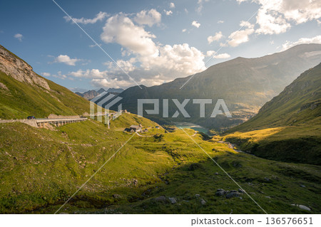 Scenic alpine mountain valley road leading toward the Pasterze Glacier with winding curves, remote mountain huts, and a turquoise glacial lake surrounded by green slopes and dramatic high Alpine peaks Scenic alpine mountain valley road leading toward the Pasterze Glacier with winding curves, remote mountain huts, and a turquoise glacial lake surrounded by green slopes and dramatic high Alpine peaks 136576651