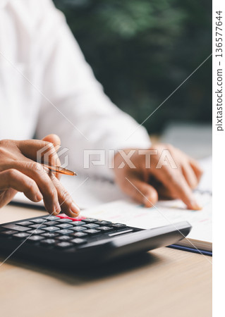 Hand writing notes in a notebook beside a calculator on office desk for budgeting and accounting. Personal finance planning, expense tracking, tax calculation, soft focus background, copy space. Hand writing notes in a notebook beside a calculator on office desk for budgeting and accounting. Personal finance planning, expense tracking, tax calculation, soft focus background, copy space. 136577644