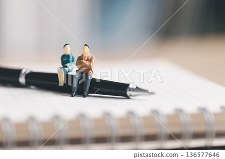 Miniature business people sitting on a pen over a notebook page, symbolizing planning and teamwork. Meeting notes, strategy, consulting and office productivity with copy space soft focus background. Miniature business people sitting on a pen over a notebook page, symbolizing planning and teamwork. Meeting notes, strategy, consulting and office productivity with copy space soft focus background. 136577646
