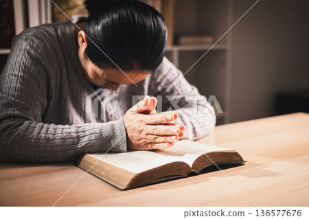 Asian woman praying with closed eyes and hands together above an open book, warm indoor light and soft focus, expressing faith, devotion, mindfulness and peaceful reflection at home. 136577676