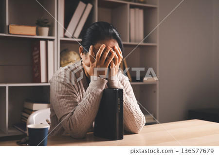 Stressed woman with hand on face beside an open Bible and glasses on a desk, warm indoor light and copy space, expressing grief, prayer, faith and seeking comfort during quiet reflection at home. Stressed woman with hand on face beside an open Bible and glasses on a desk, warm indoor light and copy space, expressing grief, prayer, faith and seeking comfort during quiet reflection at home. 136577679