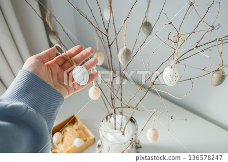 House decorating before Easter holidays: hanging small souvenir eggs on handmade Easter tree at home close up House decorating before Easter holidays: hanging small souvenir eggs on handmade Easter tree at home close up 136578247