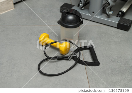 Home workout essentials: dumbbell, resistance tube, and shaker on grey tile floor 136578599