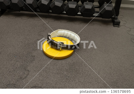 Weightlifting belt on yellow plate in gym with black dumbbells on rubber flooring Weightlifting belt on yellow plate in gym with black dumbbells on rubber flooring 136578695