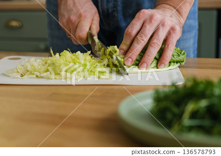 Handsome bearded man is preparing green fresh vegan salad in the kitchen at home. Male cutting vegetables, lettuce with knife on wooden chopping board, cooking vegetarian meal. Tasty healthy diet food 136578793