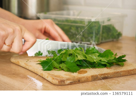 Person Chopping parsley bunch. Female cut greens with sharp knife at home kitchen. Woman hands cutting fresh aromatic herbs for cooking. Preparing food. Healthy food ingredient. Ecological products. 136579111
