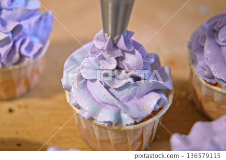 Person decorating cupcakes. Baker piping swirl of a creamy blue purple frosting onto a freshly baked cupcake. Decorated cakes with colorful sprinkles. Sweet Dessert Preparation. Pastry small business 136579115
