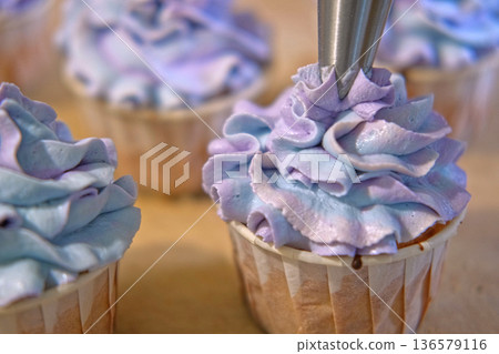 Person decorating cupcakes. Baker piping swirl of a creamy blue purple frosting onto a freshly baked cupcake. Decorated cakes with colorful sprinkles. Sweet Dessert Preparation. Pastry small business Person decorating cupcakes. Baker piping swirl of a creamy blue purple frosting onto a freshly baked cupcake. Decorated cakes with colorful sprinkles. Sweet Dessert Preparation. Pastry small business 136579116