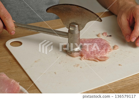 Person using meat tenderizer on raw chicken. Chef preparing pork loin slice with meat mallet on cutting board. Man tenderizing fillet. Cooking cutlets pounding poultry breast close-up. Culinary dish. 136579117