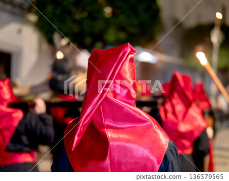 Rear view of a penitent in a red hood at a night procession 136579565