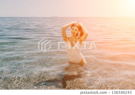 Young woman swimming, smiling in clear seawater, holding wet hair under bright summer sun. 136580243