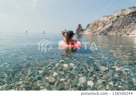 Summer vacation travel, woman swimming floating on pink ring in clear blue sea water near cliff rocks. Summer vacation travel, woman swimming floating on pink ring in clear blue sea water near cliff rocks. 136580244