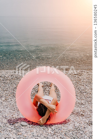 Woman floating relaxing, sunbathing with large pink inflatable ring on pebble beach vacation concept Woman floating relaxing, sunbathing with large pink inflatable ring on pebble beach vacation concept 136580245