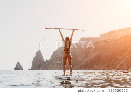 Woman Paddleboarding Ocean Coastline - A woman wearing a bikini stands on a paddleboard with her arms raised in the air, looking out at the ocean. She is surrounded by rocky cliffs. The photo is taken Woman Paddleboarding Ocean Coastline - A woman wearing a bikini stands on a paddleboard with her arms raised in the air, looking out at the ocean. She is surrounded by rocky cliffs. The photo is taken 136580252