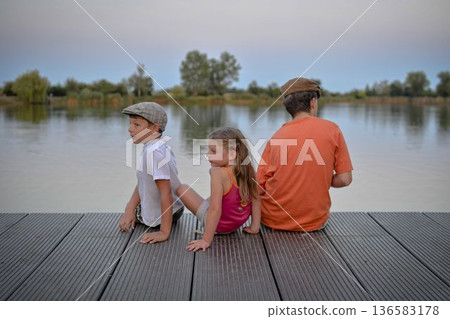 A group of young children sit on a wooden pier by a lake. The young children look toward the future. The concept of summer, holidays, and a carefree childhood. Summer and childhood concept. Children 136583178