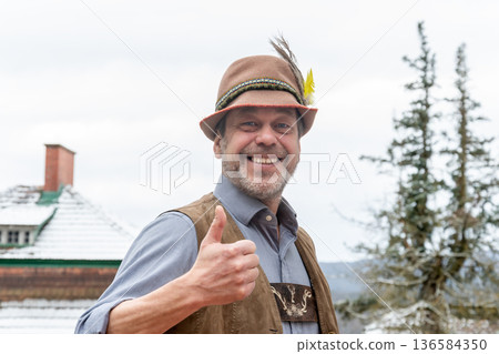 Adult man in Tyrolean hat and lederhose smiling and showing thumbs up in rural Austria Adult man in Tyrolean hat and lederhose smiling and showing thumbs up in rural Austria 136584350
