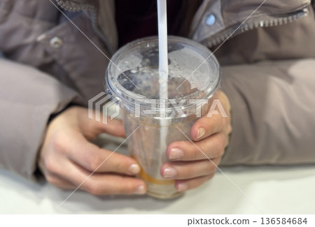 Hands Holding Iced Coffee Cup CloseUp, Student Gripping Cold Beverage With Both Hands At Cafe Table, Straw Near Lips, Condensation Beading On Plastic Cup, Casual Winter Jacket, Smartphone 136584684