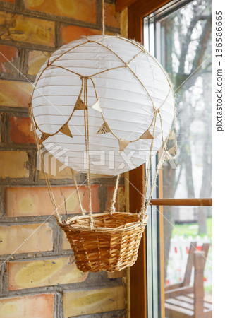 Handmade paper hot air balloon lamp with basket hanging near window in cozy room, interior decor Handmade paper hot air balloon lamp with basket hanging near window in cozy room, interior decor 136586665